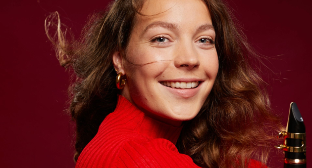 Vrouw met lang bruin haar en rode trui lacht naar de camera, houdt een klarinet vast voor een rode achtergrond