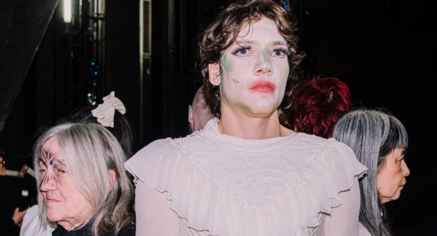 groep mensen staat in een circkel met ruggen naar elkaar toe. Twee mensen duidelijk in beeld met een geschminkt gezicht.