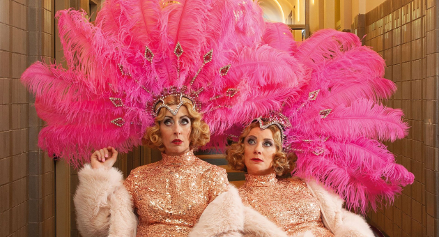 Promotiebeeld voor cabaretvoorstelling Harrekidee Plien en Bianca zitten naast elkaar en kijken bedenkelijk. Ze zijn gekleed in showgirl outfits: gouden glitterjurken, boa's en roze verentooien op hun hoofd. 