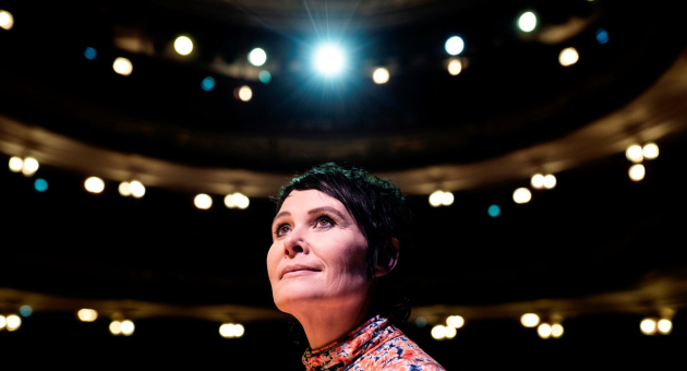 Vrouw op een toneel, belicht door een felle spot, met lege theaterstoelen op de achtergrond.