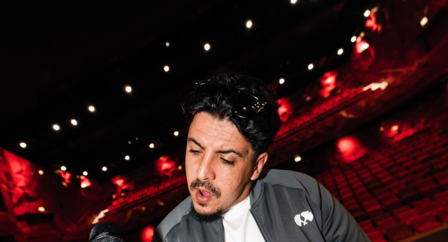 Foto van ICE op het podium. Hij draagt een zwart jack en heeft een microfoon in zijn hand. Achter hem zien we een lege theaterzaal met rode stoelen. 