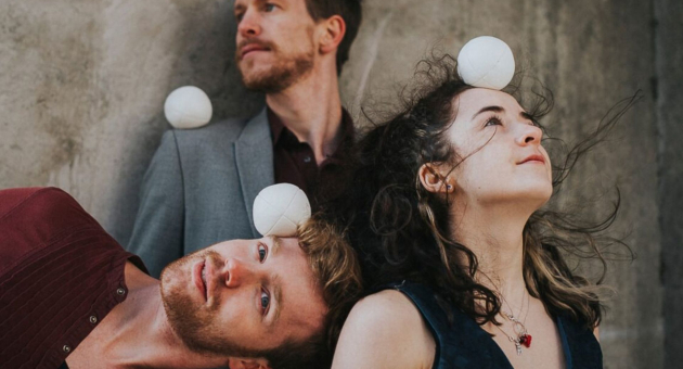 Drie mensen balanceren witte jongleerballen op hun hoofd en schouders terwijl ze dromerig omhoog kijken tegen een betonnen achtergrond.