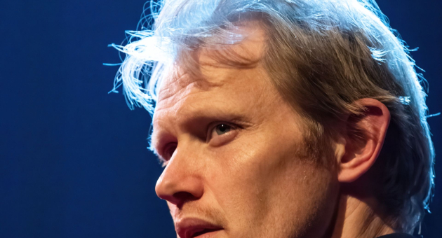 Close-up van een man met blond haar en serieuze blik, dramatisch belicht tegen een donkere achtergrond met blauw licht.