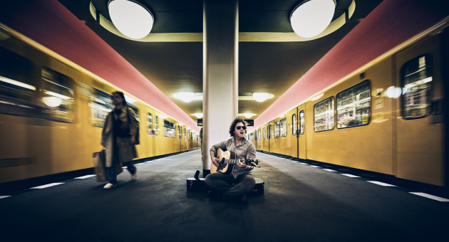 Jasper Steverlinck zittend op een station met een gitaar vast en bewegende metro.
