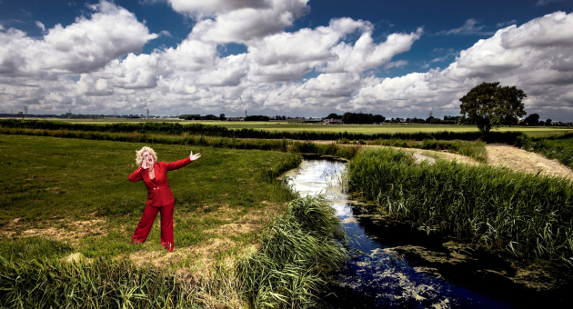 Karen Bloemen, gekleed in een rood pak, staat schreeuwend in een weiland met een blauwe wolkenlucht. 