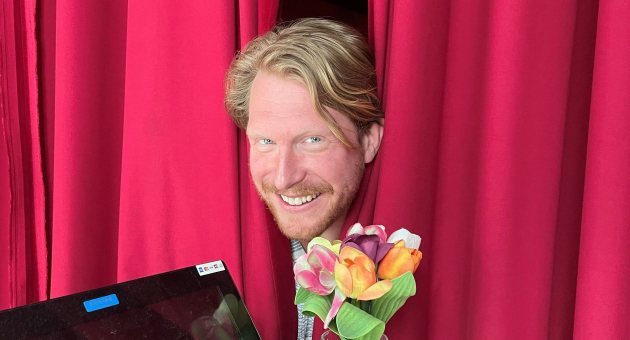 Kor Hoebe lachend tussen rode gordijnen met bloemen.