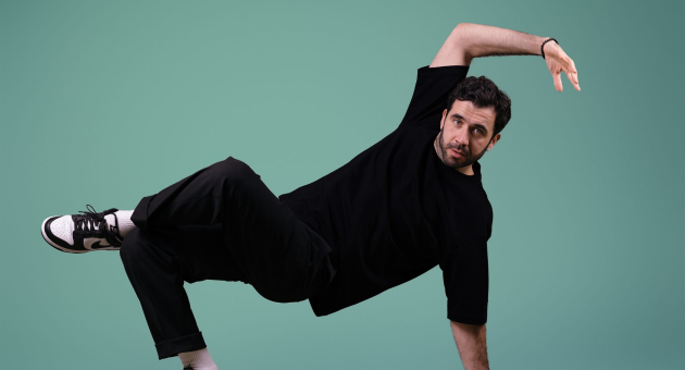 Marco Lopes gekleed in zwarte kleding in een excentrieke pose voor een lichtblauwe achtergrond. 