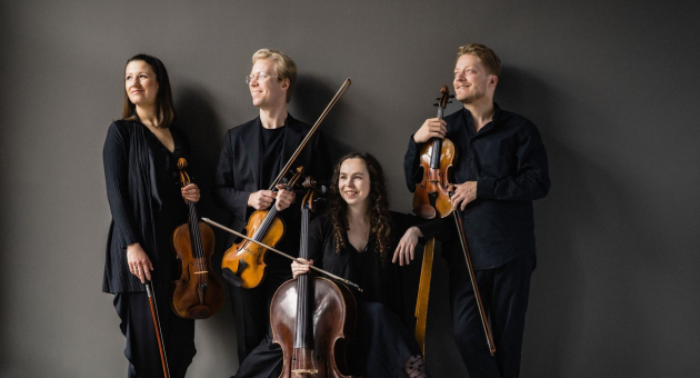 Vier musici in zwarte kleding met strijkinstrumenten poseren ontspannen tegen een grijze muur