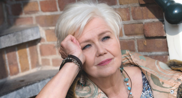 Oudere vrouw met kort wit haar leunt dromerig op haar hand, zittend bij een bakstenen muur en trap in daglicht