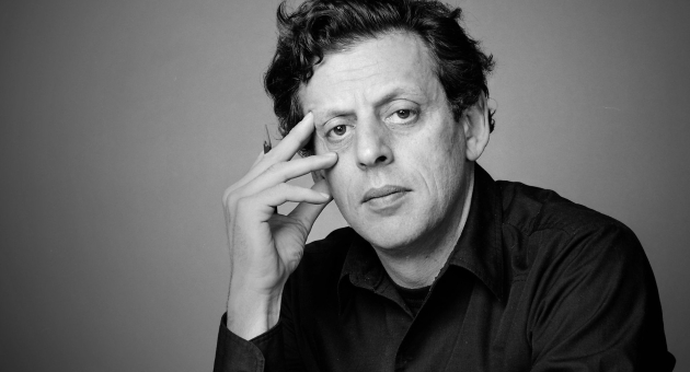 Zwart-witportret van een man met een intense blik, leunend op zijn hand, gekleed in een donker overhemd tegen een neutrale achtergrond.