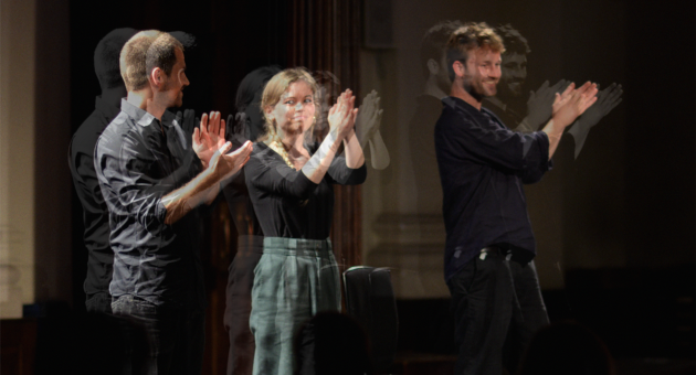 Drie mensen op een podium applaudisseren, met bewegingsonscherpte in hun handen