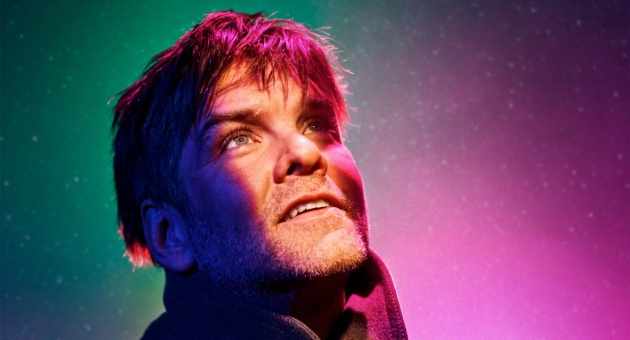 Portret van een man met warrig haar en een baard, die dromerig omhoog kijkt. Hij is kleurrijk verlicht met paarse, roze en groene tinten tegen een sterrenachtige achtergrond, wat een magische en mysterieuze sfeer creëert.