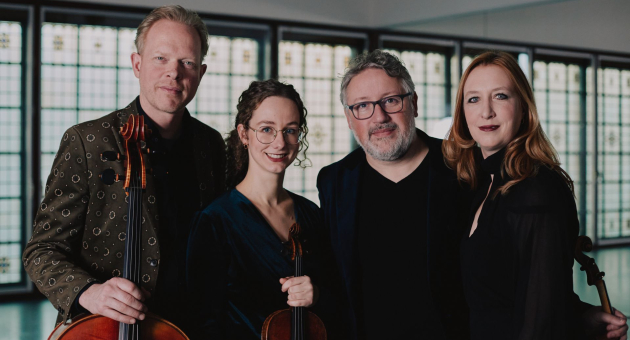 Vier mensen poseren samen in een ruimte met grote ramen met glas-in-loodpatronen. Twee van hen houden strijkinstrumenten vast. Ze kijken vriendelijk in de camera en staan dicht bij elkaar, wat een gevoel van verbondenheid uitstraalt.