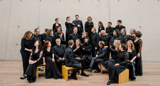 Koorleden in zwarte concertkleding poseren informeel op houten bankjes voor een strakke, betonnen wand – een speelse groepsfoto met een moderne uitstraling.