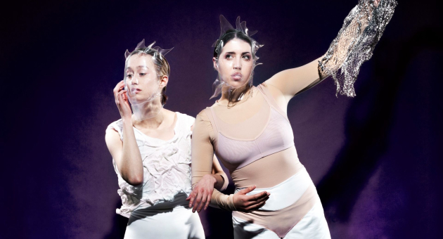 Twee performers in abstracte witte outfits en hoofdstukken poseren expressief tegen een paarse achtergrond
