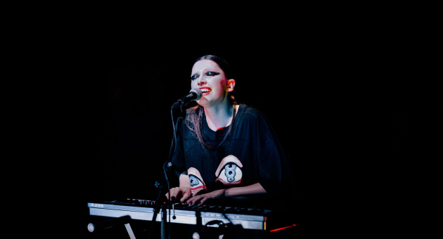  Artiest met opvallende make-up speelt keyboard en zingt op een donker podium, verlicht door een spotlight.