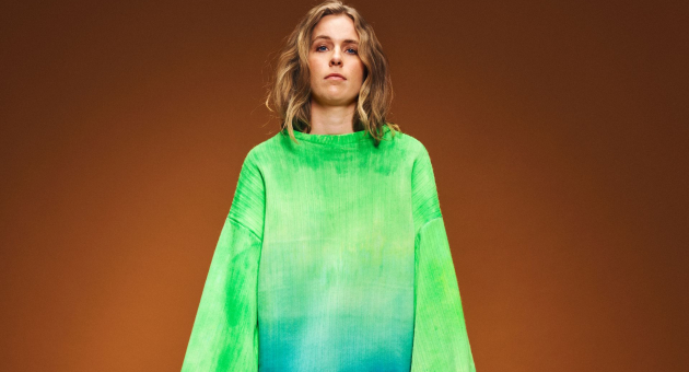Model in oversized felgroene trui met blauw kleurverloop, poseert voor een effen bruine achtergrond.