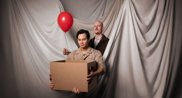 Twee mannen voor een gedrapeerd doek, één houdt een doos vast terwijl de ander een rode ballon vasthoudt.