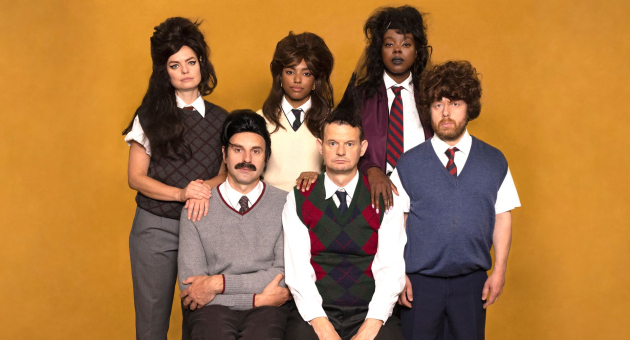 Zes personen poseren als groep tegen een okergele achtergrond, gekleed in ouderwetse outfits met pruiken, truien en stropdassen, en kijken serieus in de camera.