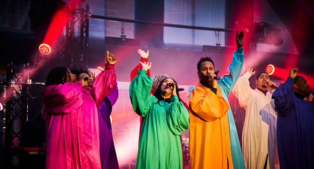 Zes zangers op podium in felgekleurde gewaden, zingend onder rood en paars podiumlicht