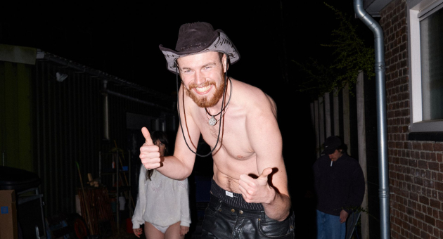 Lachende man met hoed en ontbloot bovenlichaam poseert met duimen omhoog tijdens een avondfeest.