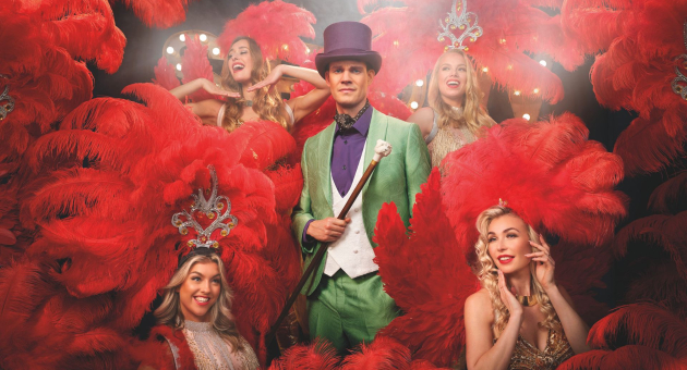 Tim Fransen is omringd door showgirls in outfits met rode veren. Tim draagt een groen pak met paarse hoed. 