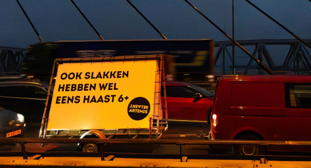 Verlichte aanhanger met tekst “Ook slakken hebben wel eens haast 6+” tussen stilstaand verkeer op brug in de avondspits