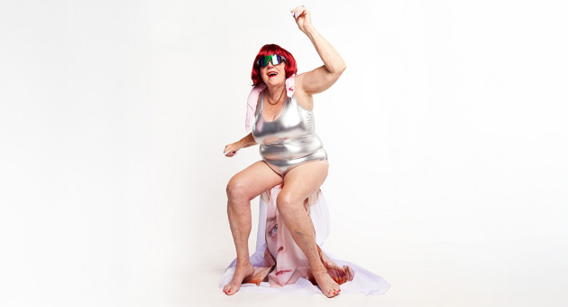 Persoon met rood haar en zonnebril in zilverkleurig badpak poseert zittend met arm omhoog op witte achtergrond