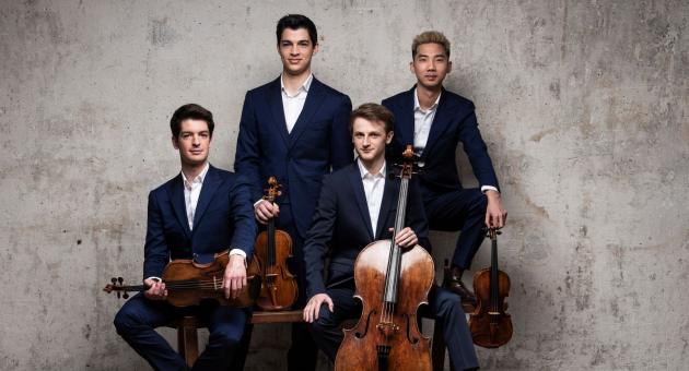 Vier mannen in donkerblauwe pakken met strijkinstrumenten voor een betonnen muur, zittend en staand in een kwartetopstelling