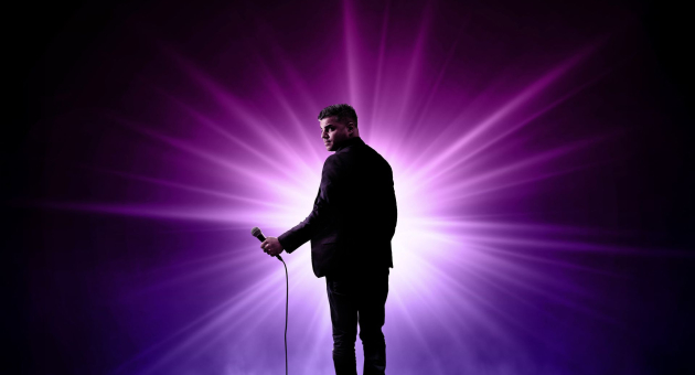 Man in pak met microfoon staat op een podium, belicht door een felle paarse lichtstraal van achteren.