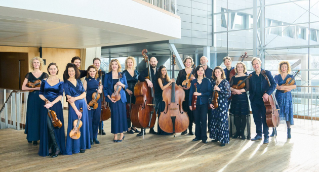 Een groep muzikanten poseert met hun strijkinstrumenten in een moderne, lichte ruimte met grote ramen. De vrouwen dragen elegante blauwe jurken, de mannen dragen zwarte pakken. Ze staan in een halve cirkel en kijken vriendelijk naar de camera.