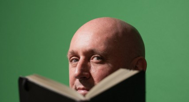 Kale man kijkt over een geopend boek richting camera, met groene achtergrond