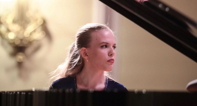Jonge vrouw speelt piano in een klassiek ingerichte ruimte met zachte verlichting. Ze kijkt geconcentreerd opzij terwijl ze speelt, met een vleugel zichtbaar op de voorgrond