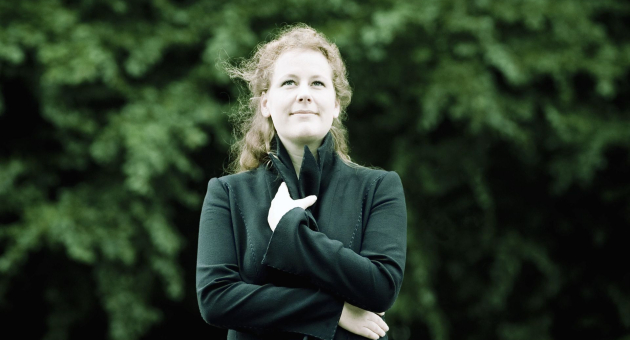 Een vrouw met rood haar staat buiten voor een groene, bladerige achtergrond. Ze draagt een donkere jas en kijkt dromerig omhoog met een lichte glimlach, terwijl ze één hand op haar borst houdt en haar armen gekruist zijn.