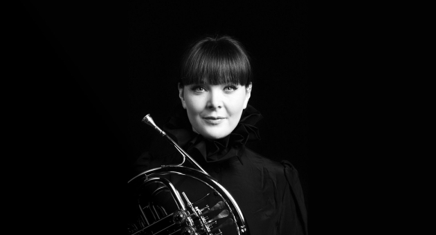 Zwart-witportret van een vrouw met een opgestoken pony en een hooggesloten, geplooide zwarte outfit. Ze kijkt rustig in de camera en houdt een hoorn (koperen blaasinstrument) vast tegen een zwarte achtergrond.