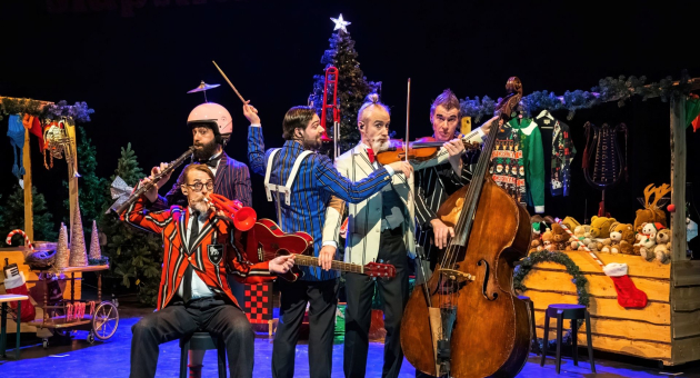 Vijf muzikanten in kleurrijke kleding bespelen verschillende instrumenten op een feestelijk versierd podium met een kerstboom en cadeaus op de achtergrond.