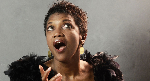 Zangeres in expressieve pose met open mond, gekleed in een zwarte outfit met veren, tegen een neutrale achtergrond.