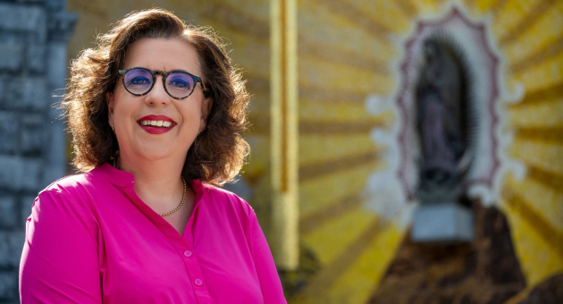 Vrouw met bril en felroze blouse lacht voor muurschildering met heiligenfiguur en stralend geel patroon