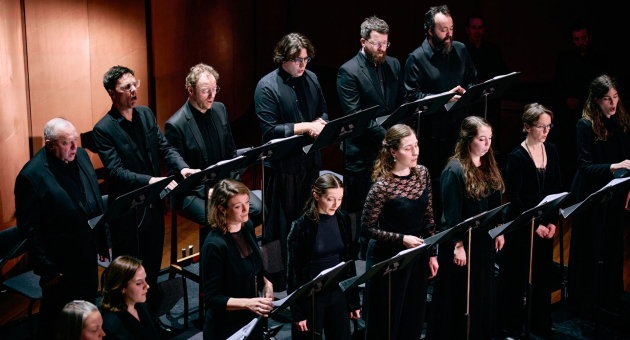 Een koor van mannen en vrouwen in zwarte kleding zingt vanaf muziekstandaards op een donker verlicht podium. Ze staan in twee rijen en concentreren zich op hun bladmuziek, met een houten wand op de achtergrond.