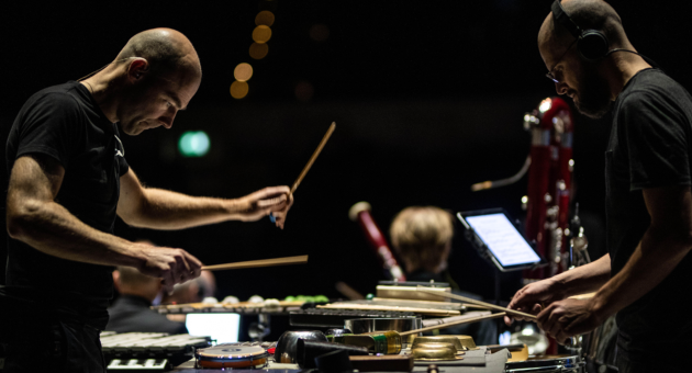  Twee mannen spelen percussie-instrumenten op een donker verlicht podium. Ze dragen zwarte kleding en zijn geconcentreerd aan het werk met drumstokken, omringd door verschillende slagwerkinstrumenten. Op de achtergrond zijn andere muzikanten vaag zichtbaar.