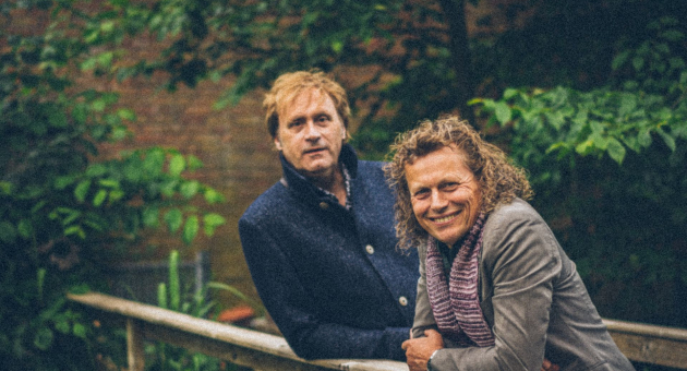 Syb van der Ploeg en Maarten Peters leunen over de reling van een brug omringt door bomen. 