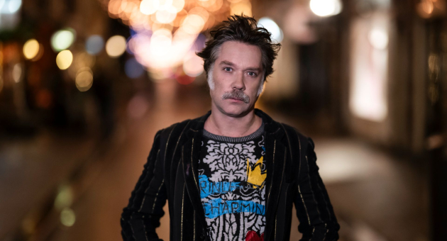 Man poseert zelfverzekerd in een sfeervol verlichte winkelstraat met onscherpe lichtjes op de achtergrond.