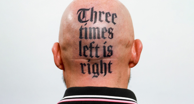 Kaalgeschoren achterhoofd van een persoon met een opvallende tatoeage in gotisch lettertype waarop staat: "Three times left is right".