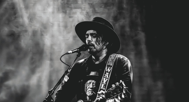 Zwart-witfoto van een man met hoed die zingt en gitaar speelt op een podium, met een expressieve blik en een shirt met een portret erop.