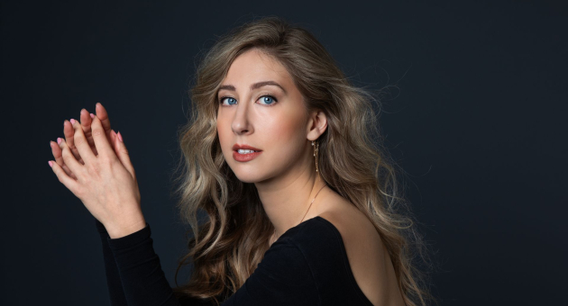  Portret van een jonge vrouw met lang, golvend blond haar tegen een donkere achtergrond. Ze draagt een elegante zwarte top met open rug en kijkt met een rustige, zelfverzekerde blik in de camera, terwijl ze haar handen gevouwen houdt.