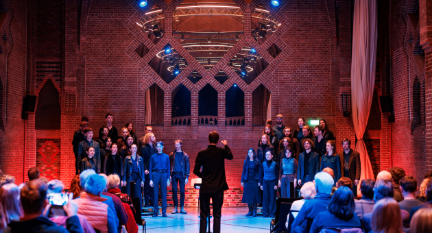 Het Nederlands Studenten Kamerkoor voert een concert uit in een sfeervol verlichte kerk, onder leiding van een dirigent.