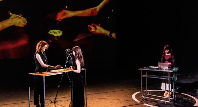 Drie performers op een donker podium tijdens een multimediale voorstelling met live projecties.