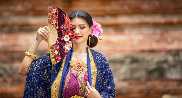 Traditioneel geklede Aziatische vrouw met waaier en bloemen in het haar poseert voor een historische muur.