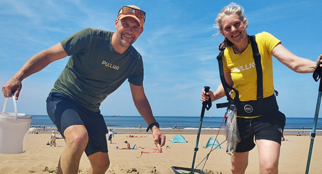 Twee mensen verzamelen afval op het strand met een emmer en grijper, op de achtergrond zee en strandtenten.