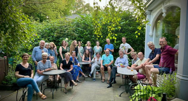 Groep van ruim twintig mensen op een terras omringd door groen, zittend en staand in informele setting